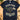 A navy blue graphic t-shirt laid flat on a wooden desk, surrounded by lifestyle props including a potted plant, a coffee mug, eyeglasses, a notebook, and a stethoscope. The t-shirt features a graphic of a cloud wearing sunglasses with lightning bolts and text reading 'BLACK CLOUD SOCIAL CLUB' and 'LIFETIME MEMBER'.