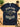 A navy blue graphic t-shirt laid flat on a wooden desk, surrounded by lifestyle props including a potted plant, a coffee mug, eyeglasses, a notebook, and a stethoscope. The t-shirt features a graphic of a cloud wearing sunglasses with lightning bolts and text reading 'BLACK CLOUD SOCIAL CLUB' and 'LIFETIME MEMBER'.