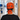 A person viewed from behind wearing a bright orange surgical scrub cap with a black ribbon tie, featuring a Halloween cat print and the text 'I FOUND THIS HUMERUS'. The background shows a blurred hospital hallway.